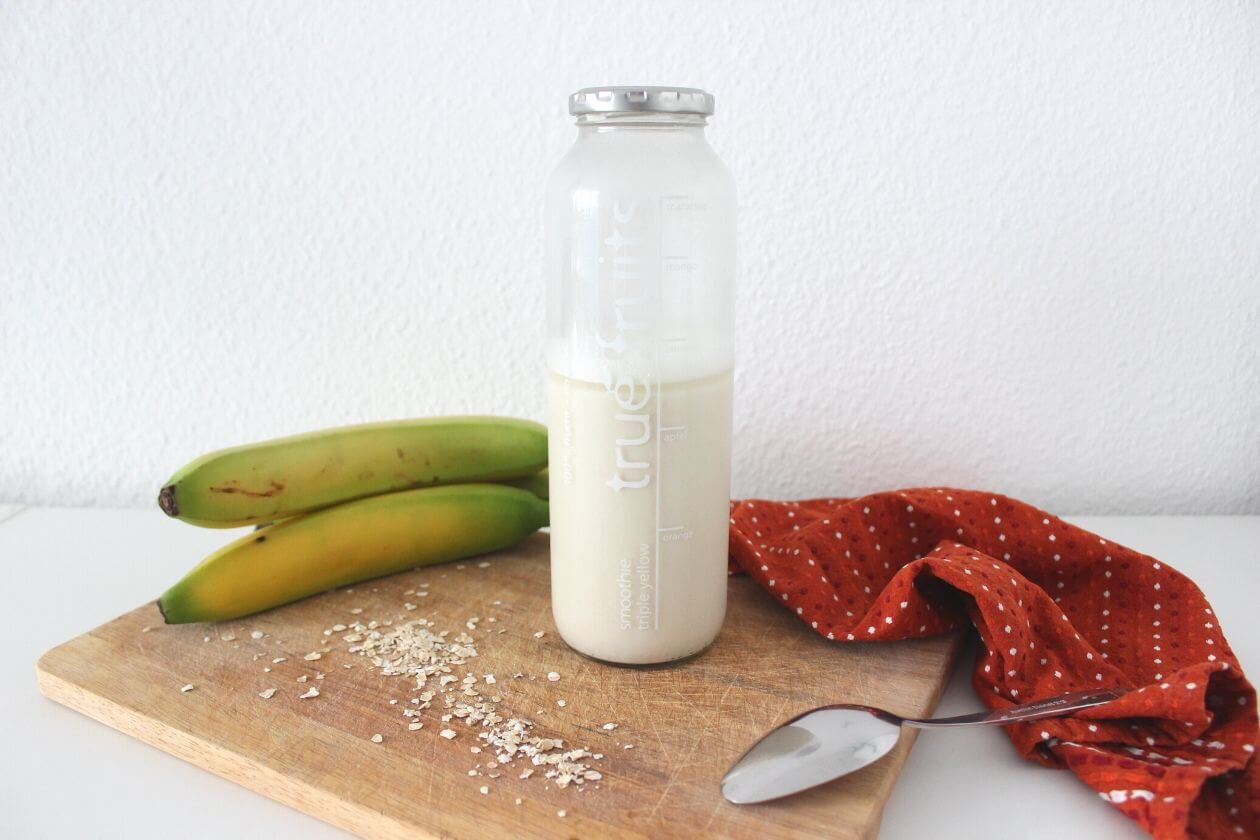 Ein Flasche Hafermilch auf einem Brett, im Hintergund Bananen und eine rote Stoffserviette